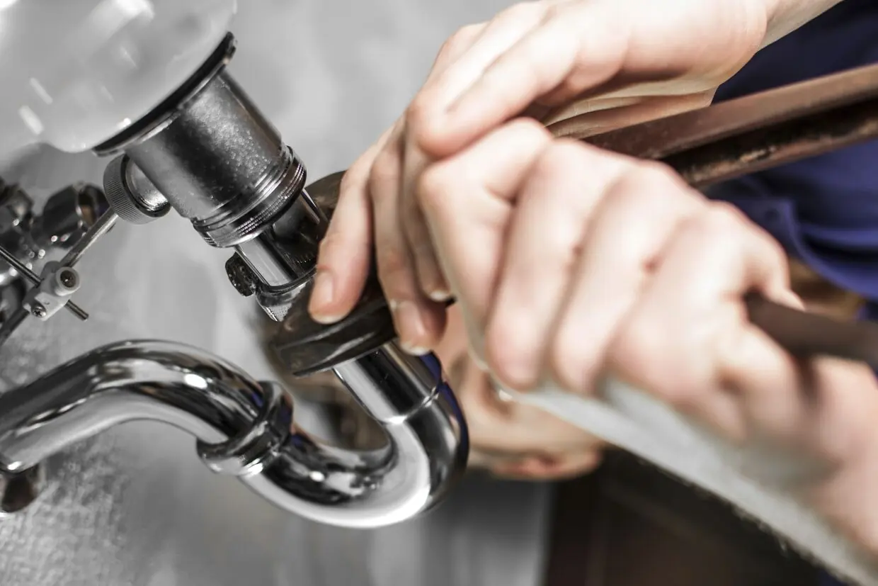 Monteur löst mit dem Schlüssel die Überwurfmutter am Siphon unter einem Waschbecken.