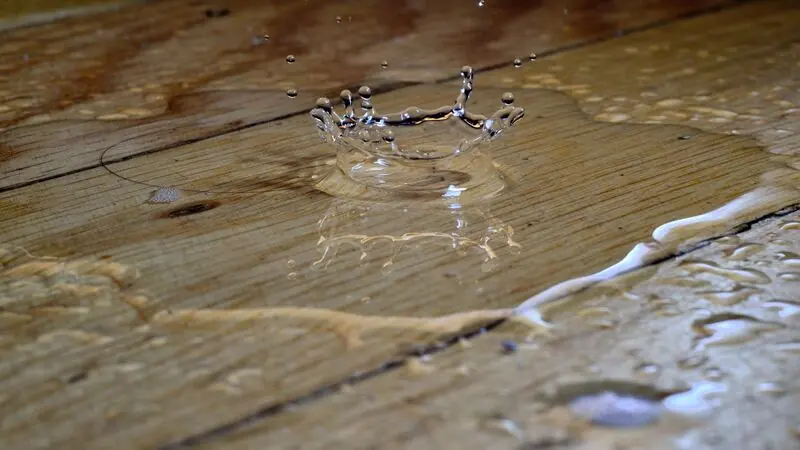 Ausgetretenes Wasser steht auf einer Holzdiele, ein Tropfen schlägt in der Pfütze auf.